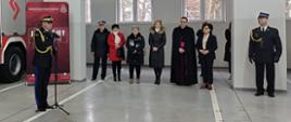 Zastępcą komendanta głównego PSP przemawiający przez mikrofon. W tle pozostali uczestnicy uroczystości. 