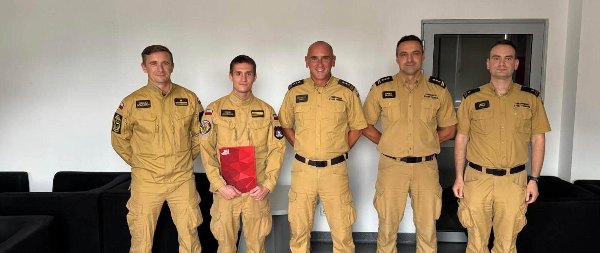 Grupowe zdjęcie pięciu funkcjonariuszy Państwowej Straży Pożarnej w mundurach. Drugi od lewej trzyma czerwoną teczkę podczas uroczystego spotkania w pomieszczeniu służbowym.