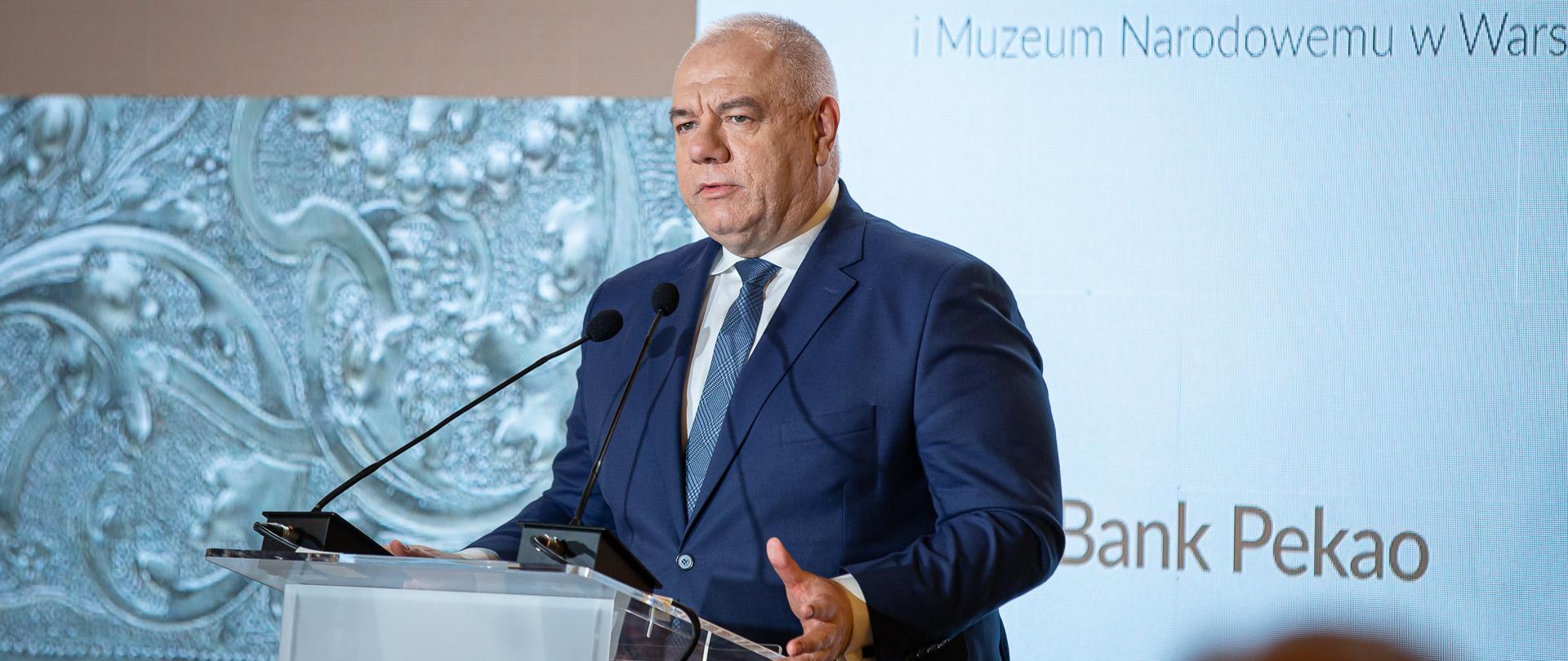 Wicepremier Jacek Sasin stoi przy mównicy, bliski plan. W tle ścianka z napisem Bank Pekao S.A. 