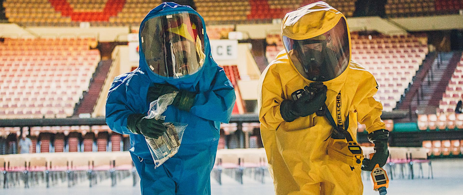 Dwaj strażacy specjalistycznej grupy ratownictwa chemiczno-ekologicznego ubrani w kombinezony ochrony przeciwchemicznej w kolorze niebieskim i żółtym podczas akcji. 