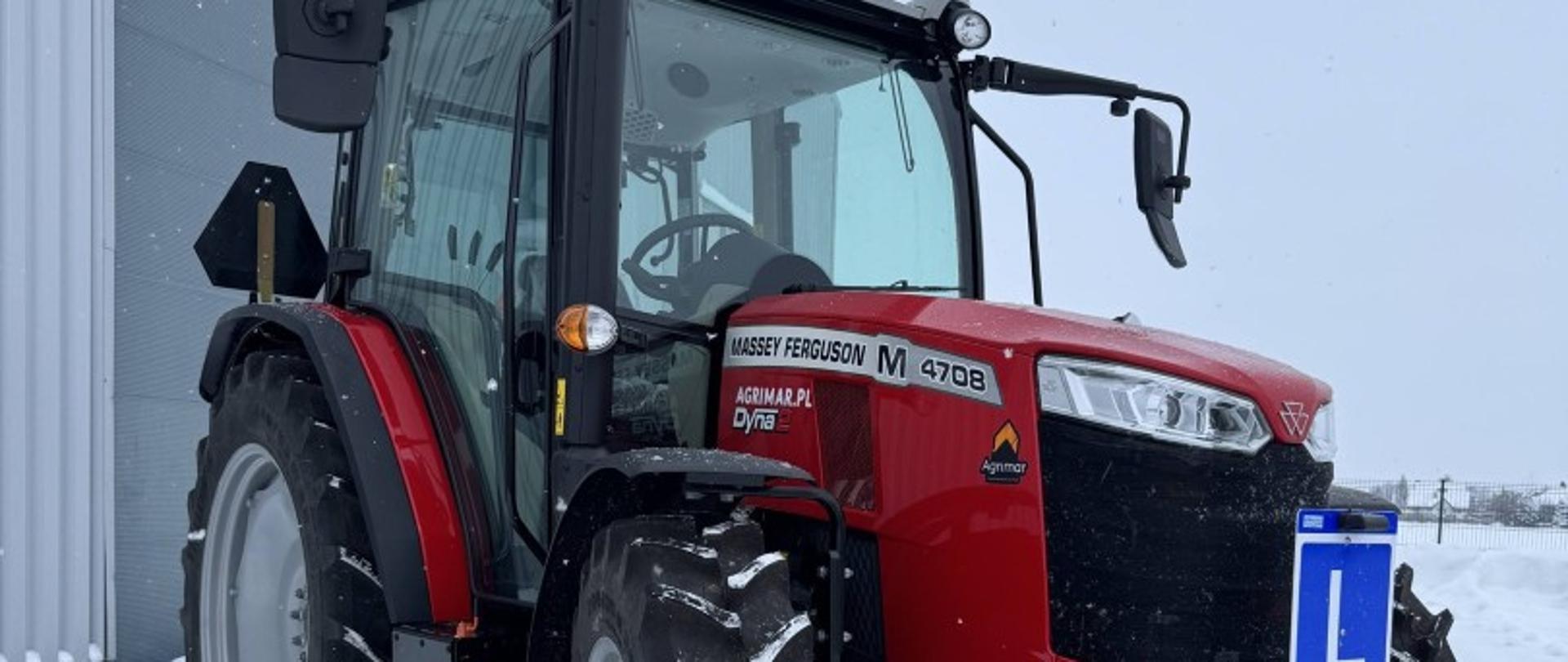 Czerwony ciągnik rolniczy Massey Ferguson zaparkowany na zaśnieżonym placu przy hali. Maszyna szkoleniowa przygotowana do nauki jazdy.