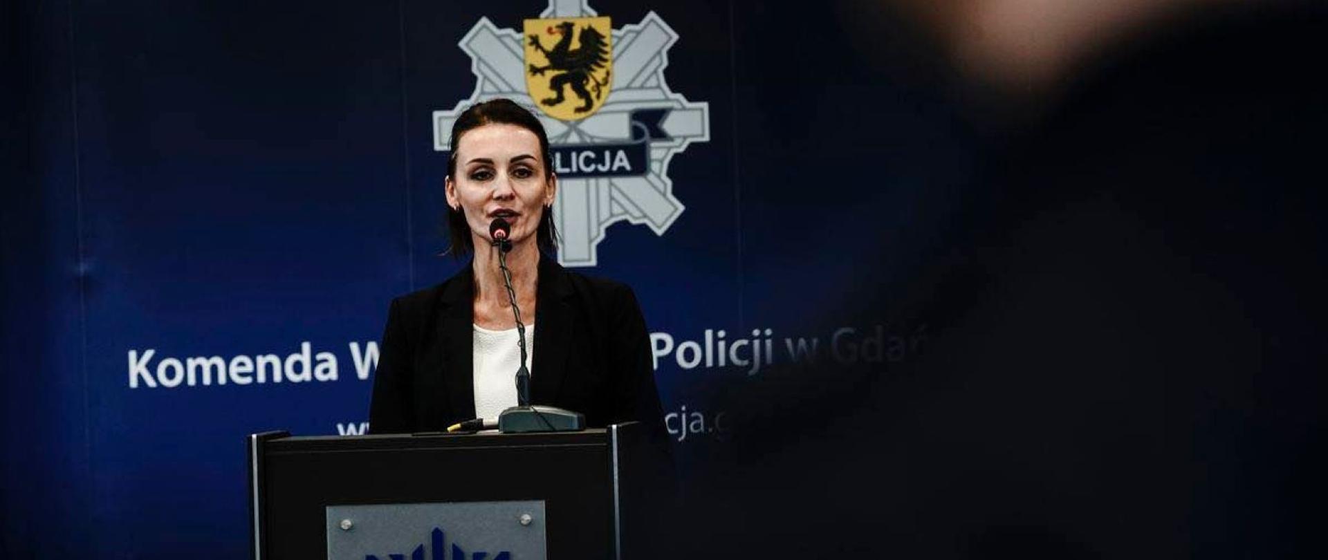Kobieta (wojewoda) stoi przy mównicy, za nią napis Komenda Wojewódzka Policji w Gdansku