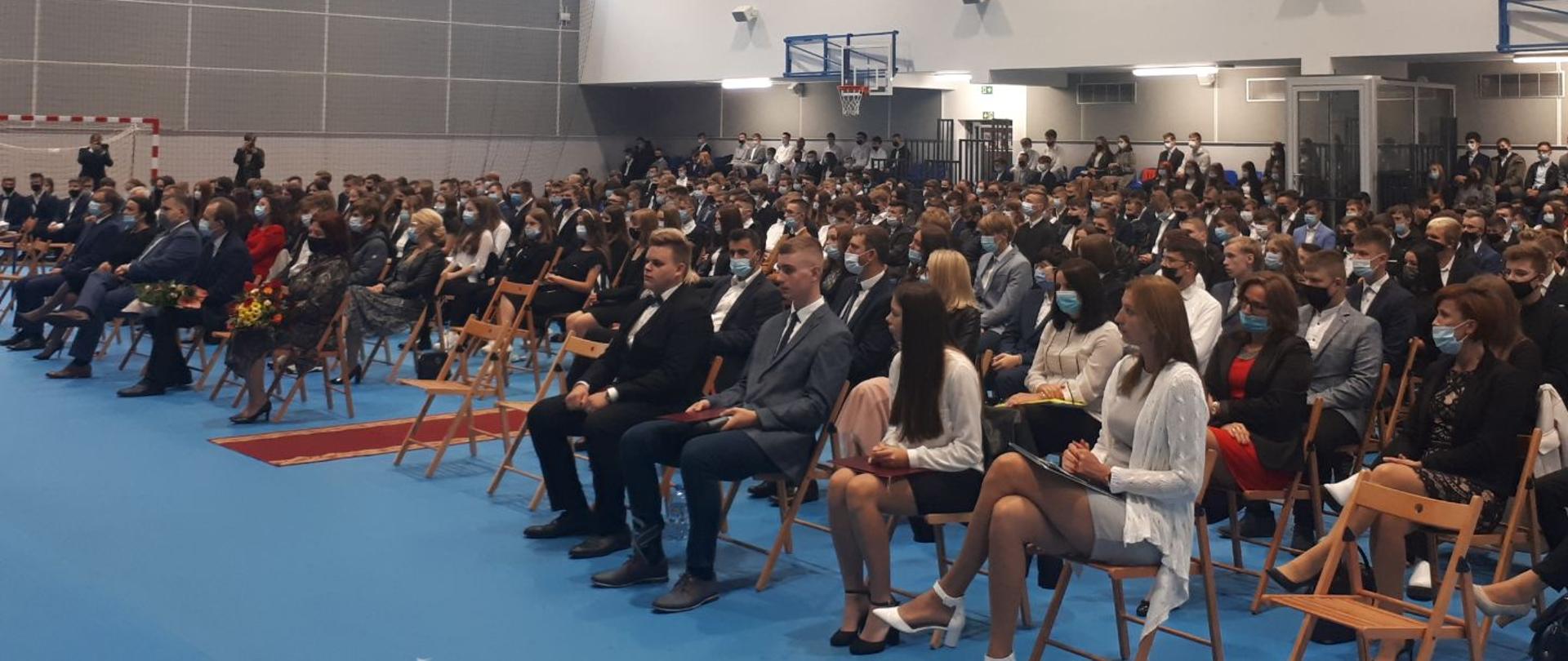 Duża sala gimnastyczna z niebieską podłogą. Na krzesłach siedzą uczniowie i nauczyciele. Są uroczyście ubrani. W tle kosz do piłki siatkowej. 