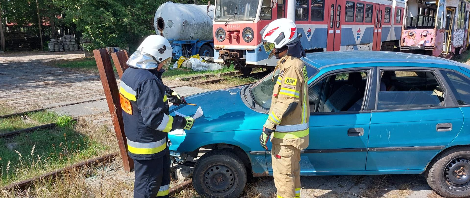 Dwóch druhów ochotniczych straży pożarnych podczas zajęć kierującego działaniem ratowniczym dla strażaka- ratownika OSP. Dowodzący akcją wskazuje miejsce cięcia na samochodzie drugiemu druhowi. 