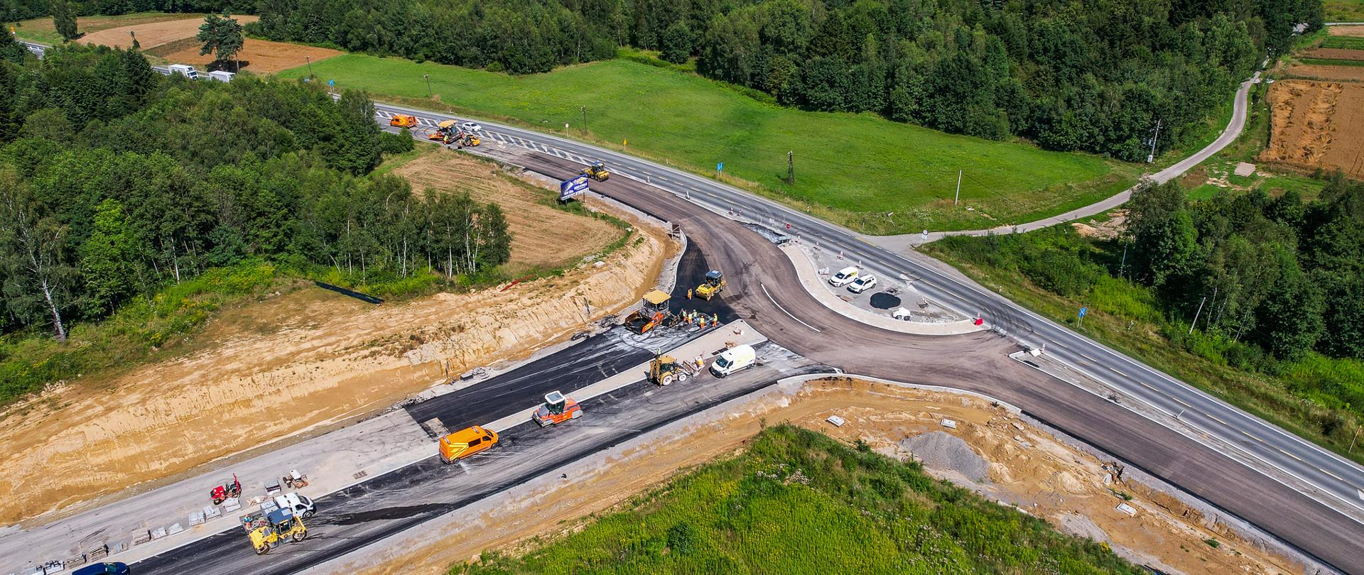 Zdjęcie lotnicze przedstawiające budowę ronda na skrzyżowaniu dwóch dróg. Widać na budowanym odcinku pojazdy z budowy, na drodze widać samochody. Widać zarys ronda. jest pogodnie świeci słońce.