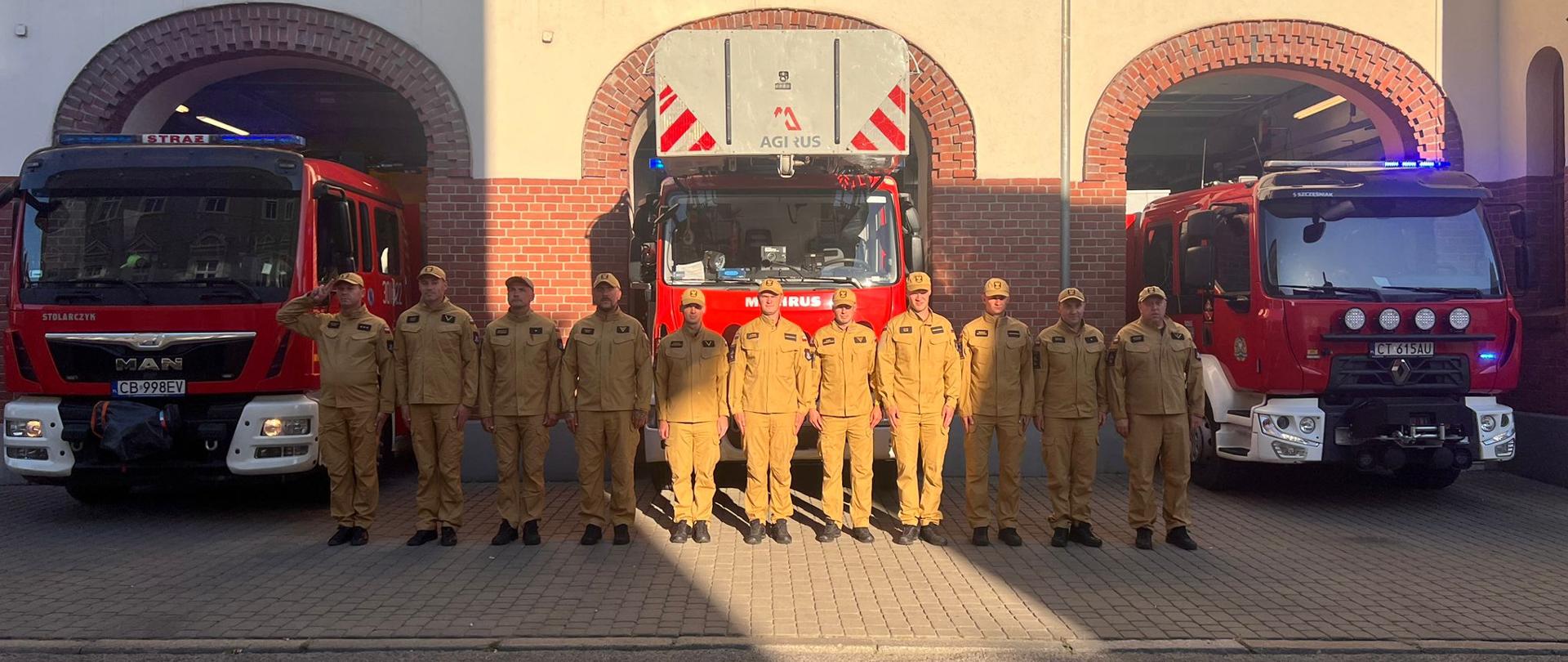 Zbiórka przed budynkiem JRG1 w Bydgoszczy.Strażacy w szeregu w żółtych ubraniach służbowych. Za nimi trzy pojazdy pożarnicze czerwone. 