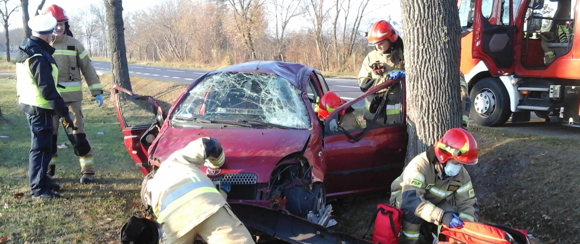 Uszkodzony samochód osobowy Toyota Yaris na poboczu drogi. Przy samochodzie pracujący strażacy. W tle stojący na jezdni samochód ratowniczo-gaśniczy.