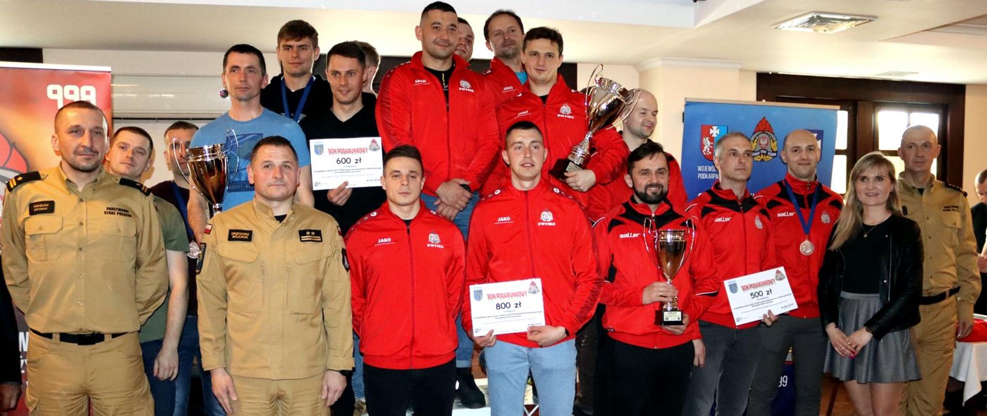 Zdjęcie zrobione w pomieszczeniu, podczas wręczenia nagród po zawodach sportowych. Zdjęcie grupowe zawodników oraz osób zaproszonych na zawody. Trzech zawodników trzyma w dłoniach promesy nagród finansowych. 