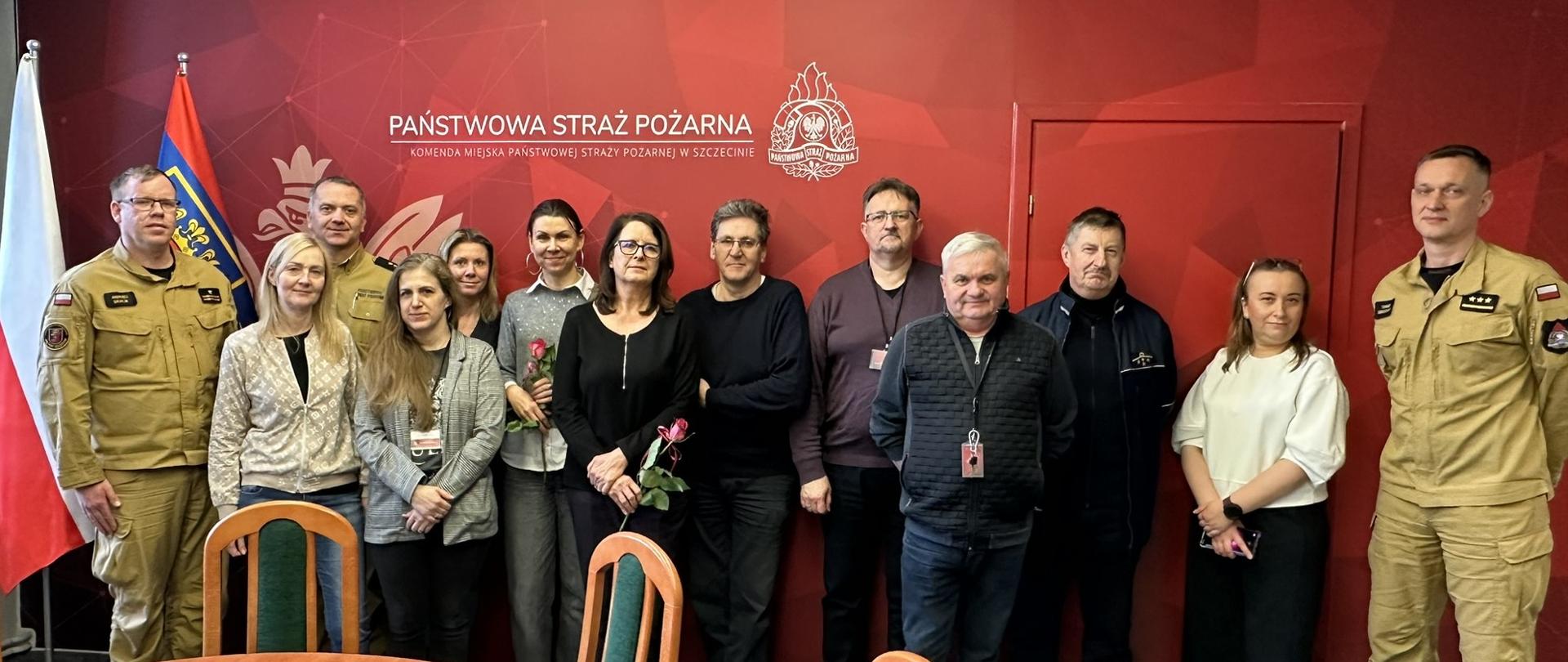 Na zdjęciu widać grupę osób stojących razem w pomieszczeniu z czerwonym tłem, na którym widnieje napis „Państwowa Straż Pożarna” oraz logo straży pożarnej. Część osób ubrana jest w mundury strażackie, a pozostali w ubrania cywilne. Niektóre kobiety trzymają w rękach czerwone róże. W tle widoczna jest również flaga Polski i flaga Państwowej Straży Pożarnej. Zdjęcie wygląda na pamiątkowe, wykonane podczas spotkania lub uroczystości w siedzibie straży pożarnej.