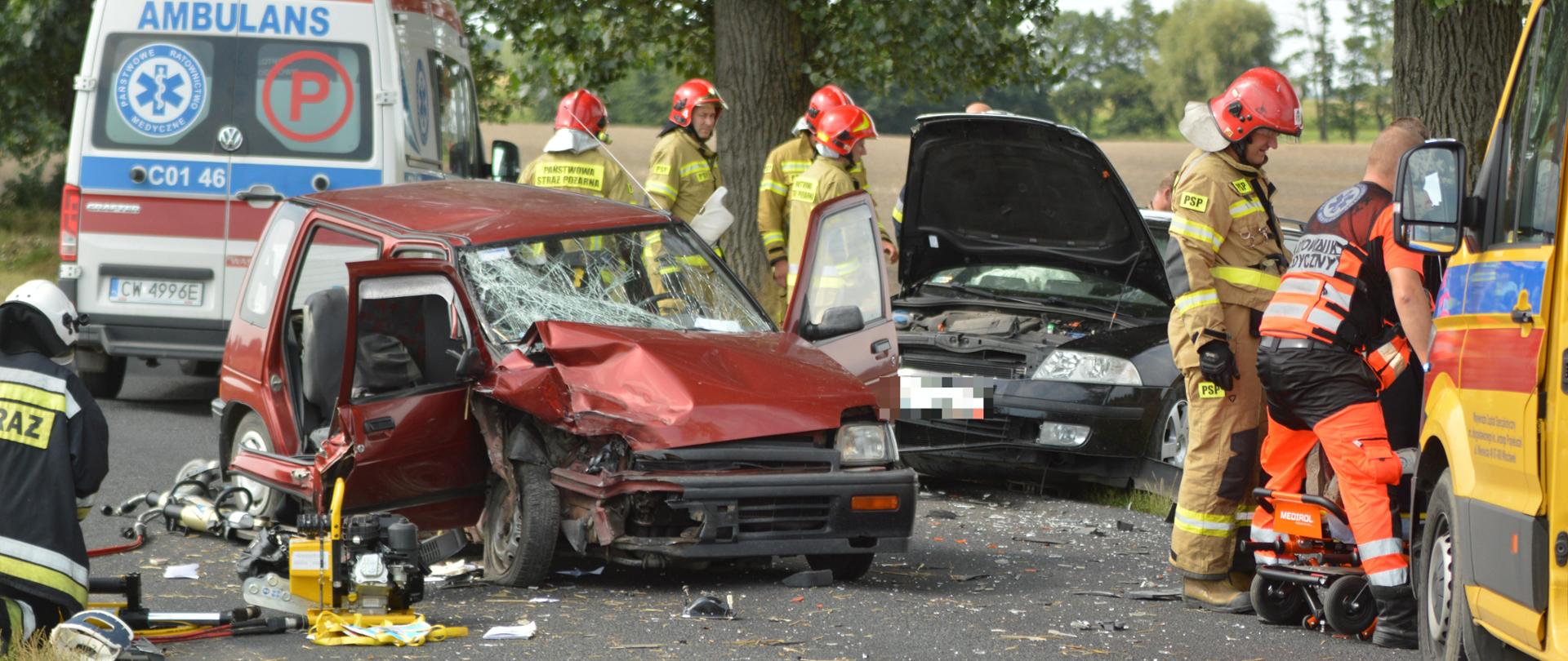 Na zdjęciu widać dwa rozbite auta osobowe. Wokół pracują strażacy w liczbie 5 z PSP, 1 z OSP oraz służba lotniczego pogotowia ratunkowego. Z prawej strony widać fragment kabiny karetki pogotowia, za wrakami stoi tyłem ambulans. W tle drzewa.