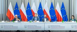 Kierownictwo MSWiA siedzi przy stole prezydialnym. W tle flagi polskie i unijne 