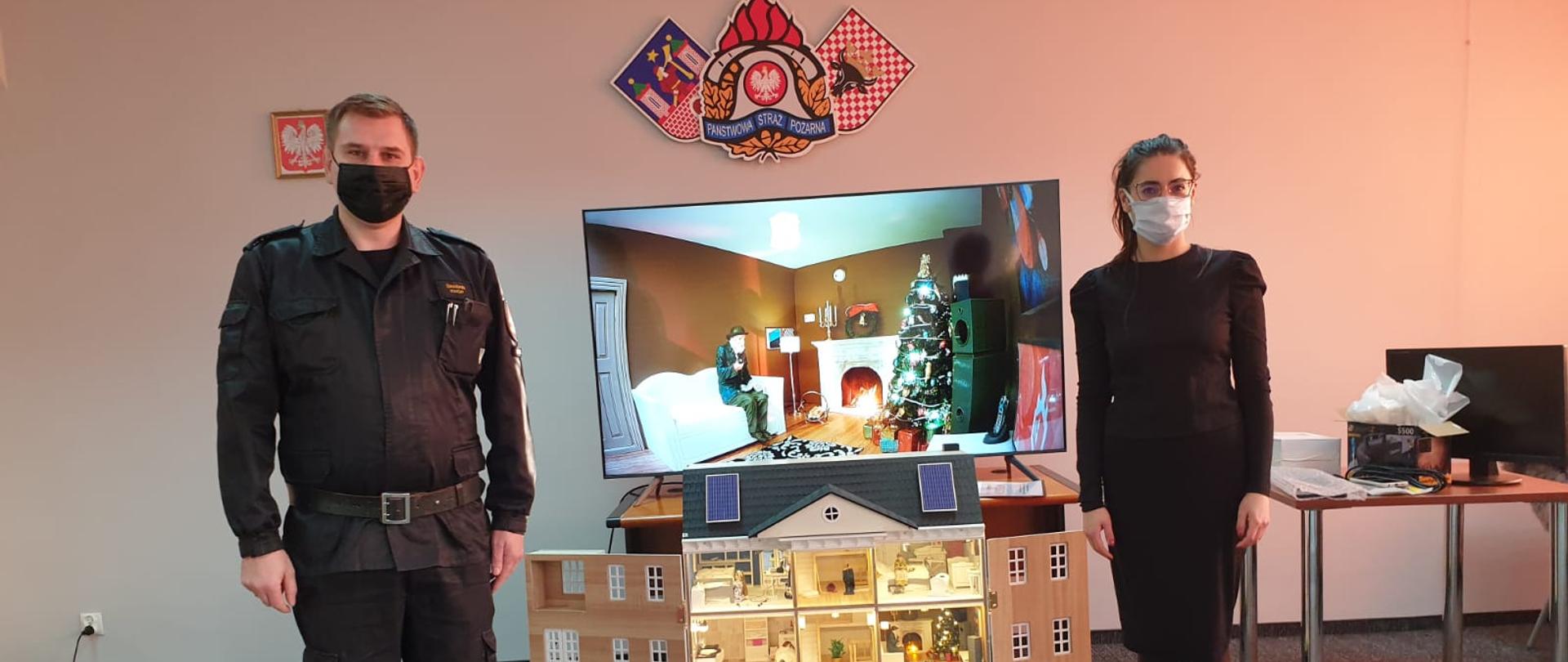 Na zdjęciu widać w centralnym punkcie mobilny symulator zagrożeń domku jednorodzinnego. Na poszczególnych piętrach pomieszczenia ze standardowym wyposażeniem domu jednorodzinnego. Na dole znak Wojewódzkiego Funduszu Ochrony Środowiska i Gospodarki Wodnej. Za domkiem znak Państwowej Straży Pożarnej i godło powiatu. Po bokach domku widać Zastępcę Komendanta Miejskiego Państwowej Straży Pożarnej w Kaliszu kpt. Damiana Knopa oraz przedstawicielkę firmy Mlabs z Poznania Panią Mariannę Krzakiewicz