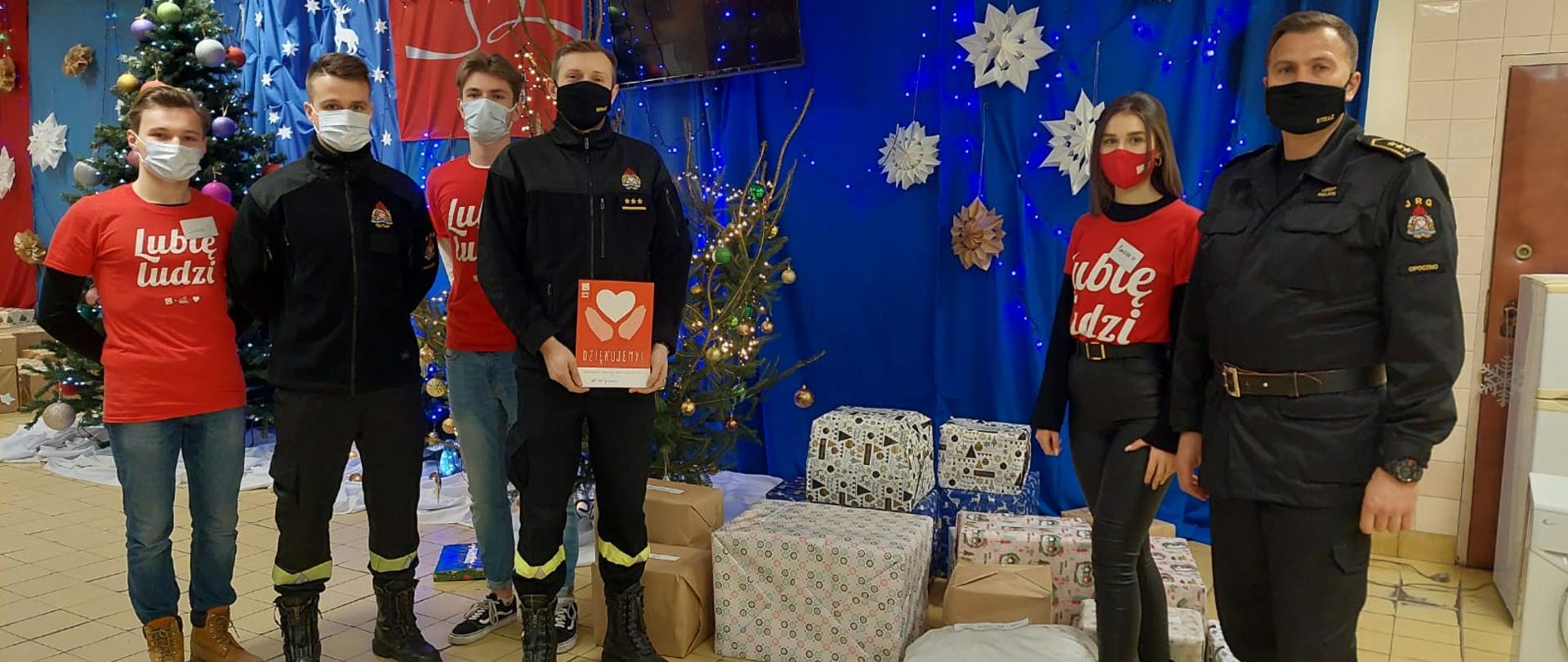 Troje strażaków w ubraniach koszarowych, jeden strażak trzyma czerwony dyplom. Pośród nich troje wolontariuszy szlachetnej paczki ubranych w czerwone koszulki z napisem lubię ludzi. Wszyscy zgromadzeni mają założone maseczki. W tle zapakowane prezenty oraz dekoracje świąteczne i choinki na niebieskim tle. 