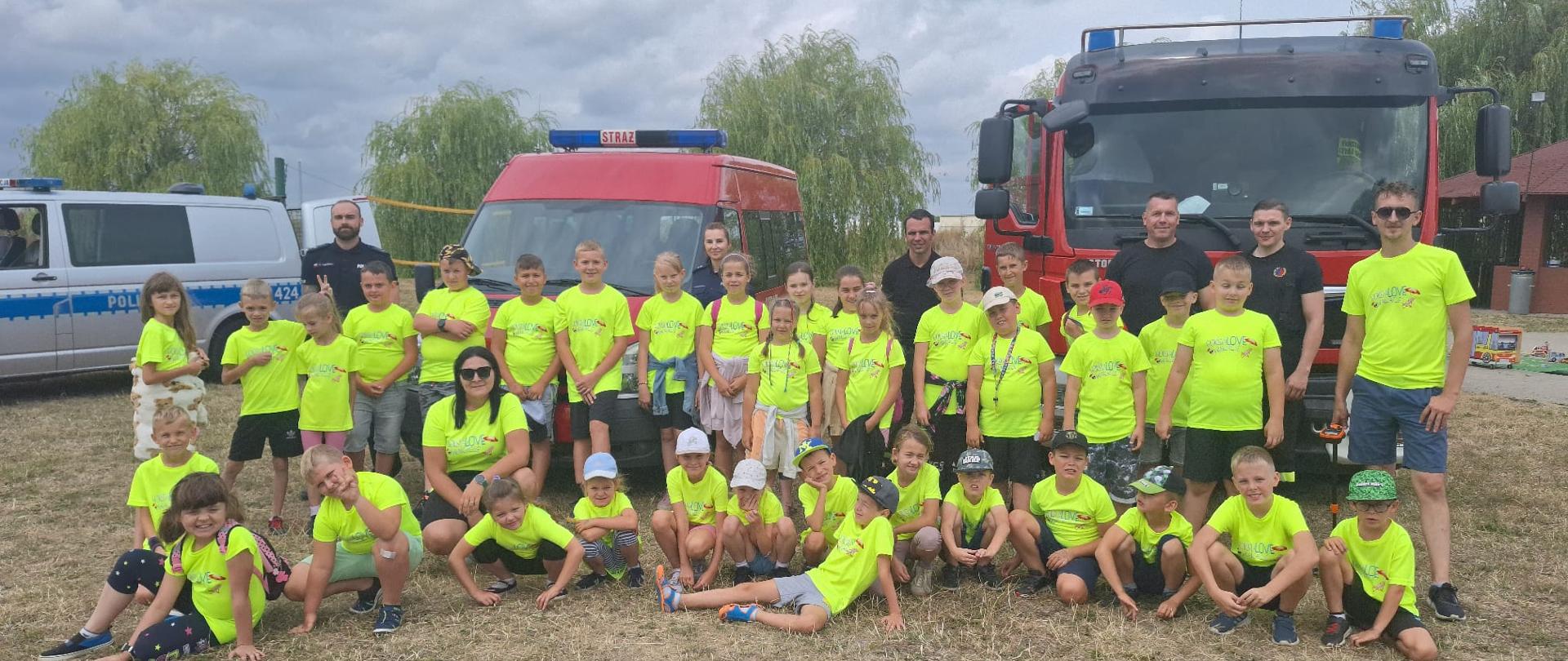Zdjęcie przedstawia strażaków, policjantów oraz grupę dzieci pozujących do zdjęcia. W tle wozy strażackie oraz radiowóz policyjny. Dzieci ubrane są w jednakowe koszulki