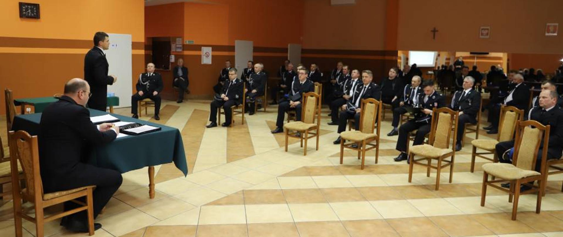 Zdjęcie przedstawia funkcjonariuszy Komendy Powiatowej Państwowej Straży Pożarnej w Zambrowie oraz druhów Ochotniczej Straży Pożarnej ubranych w mundury wyjściowe podczas spotkania w sprawie ustawy o ochotniczej straży pożarnej, w budynku Gminnego Ośrodka Kultury w Szumowie.