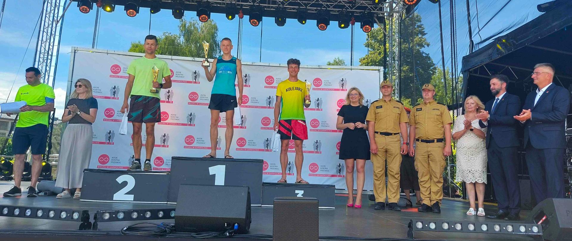 Na scenie na podium zawodnicy, obok nich stoją oficjalni goście