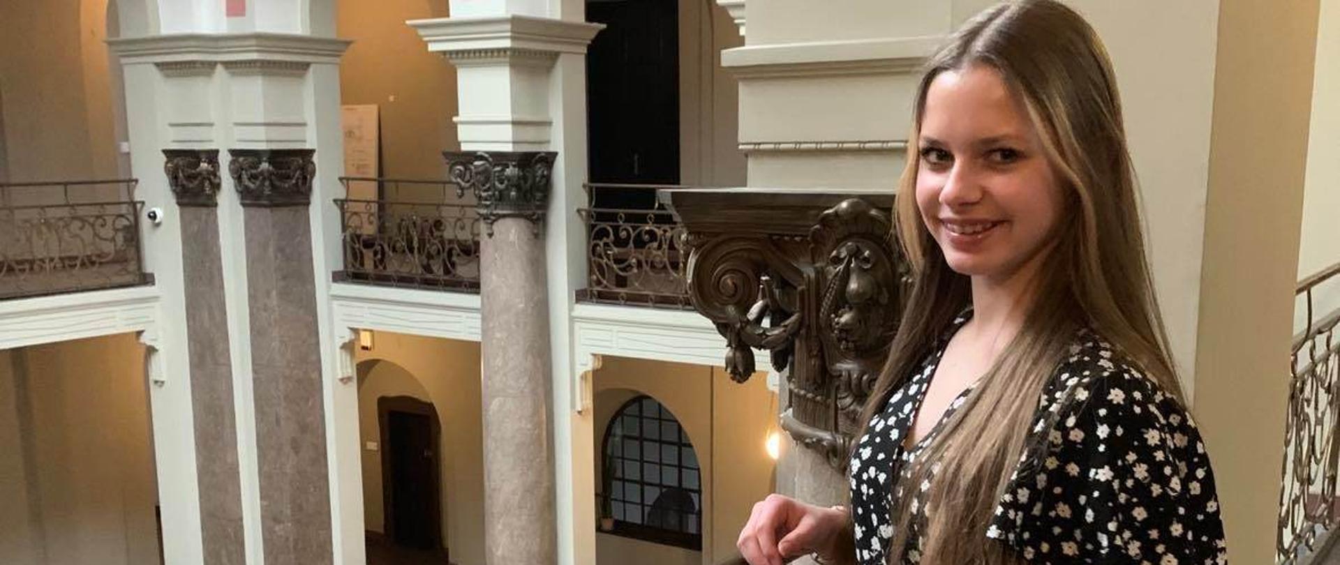 Dziewczyna, długie włosy, sukienka czarna w białe plamki, na tle dziedzińca stoi przy balustradzie