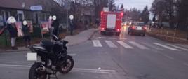 Widać motocykl na drodze, po lewej osoby postronne, z przodu samochód strażacki