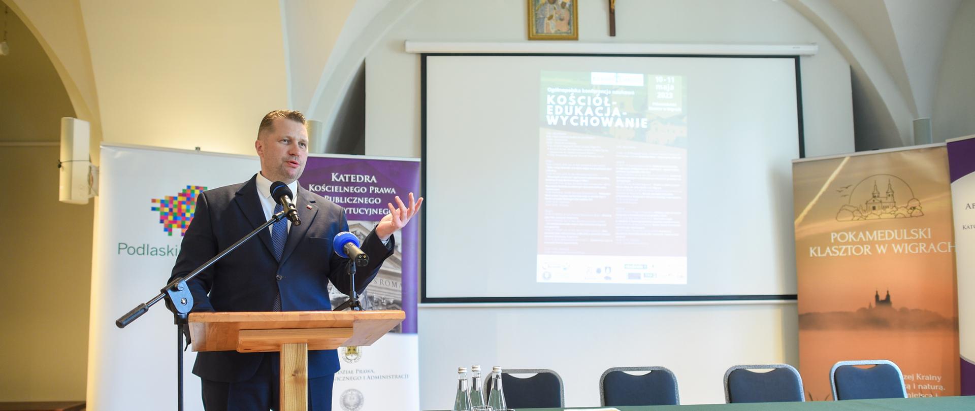 Za drewnianą mównicą stoi minister Czarnek i mówi do mikrofonu, obok niego stół nakryty zielonym materiałem, za nim na białej ścianie wisi wielki ekran, nad nim krzyż.