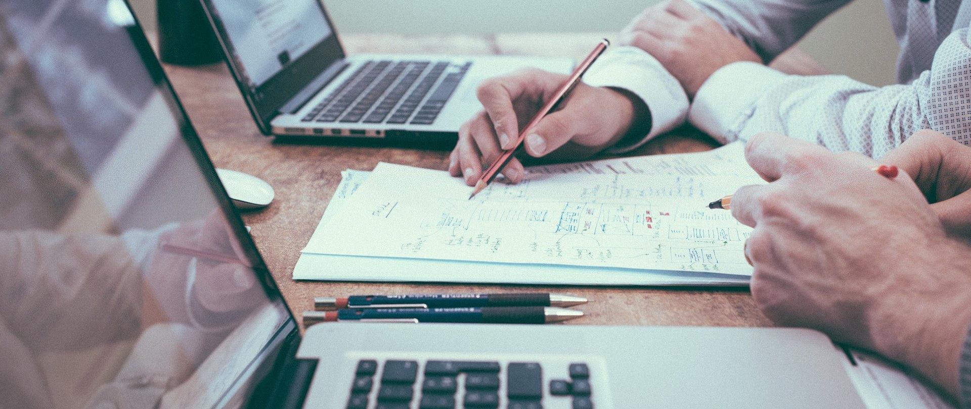 Zdjęcie przedstawia dwa laptopy, widziane z boku. Obok ręce dwóch postaci, trzymające długopisy i piszące niewyraźne notatki na kartce.