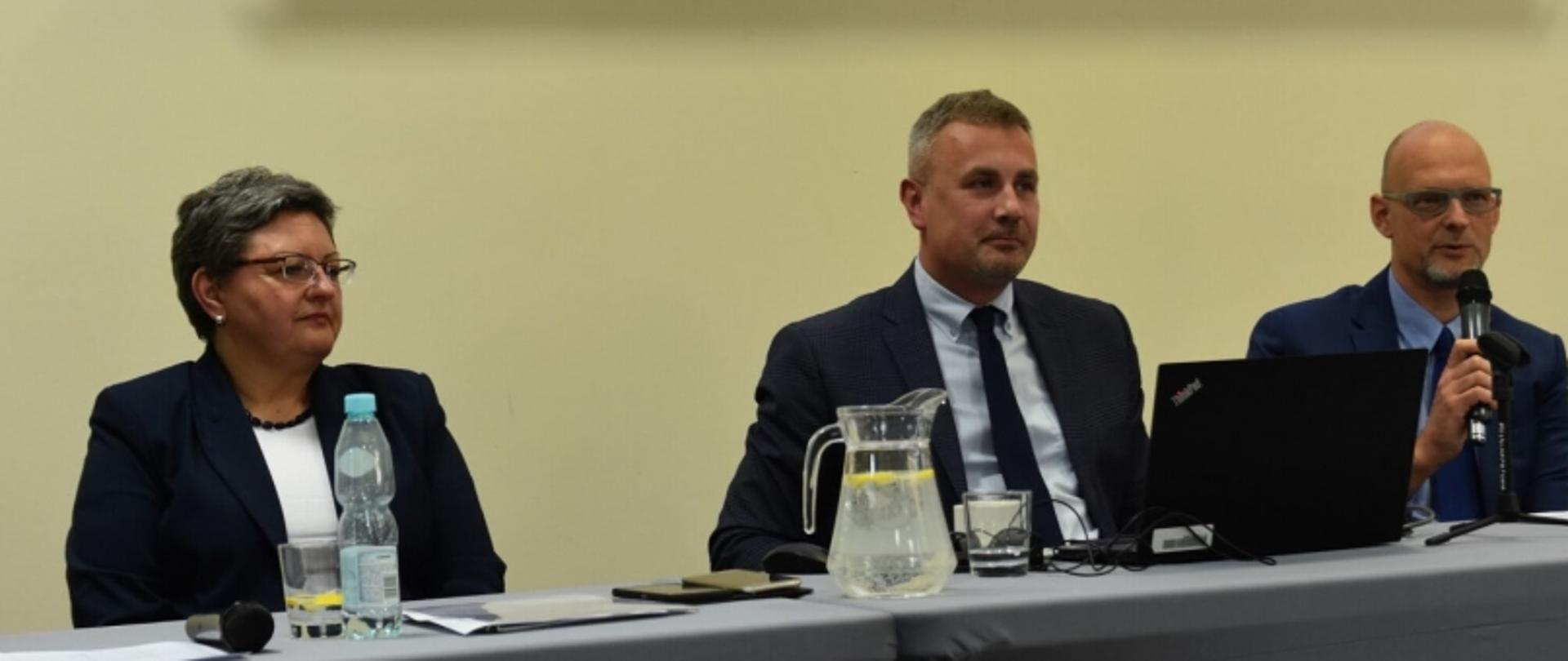 Trzy osoby przy stole konferencyjnym z dokumentami, wodą, mikrofonem i laptopem.