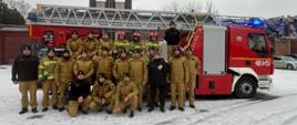 Grupa strażaków biorących udział w szkoleniu kierowców‑operatorów drabiny mechanicznej pozuje przed samochodem z drabiną Rosenbauer. Zdjęcie wykonano na placu KP PSP w Prudniku, w zimowych warunkach, na tle wieży ćwiczeń.