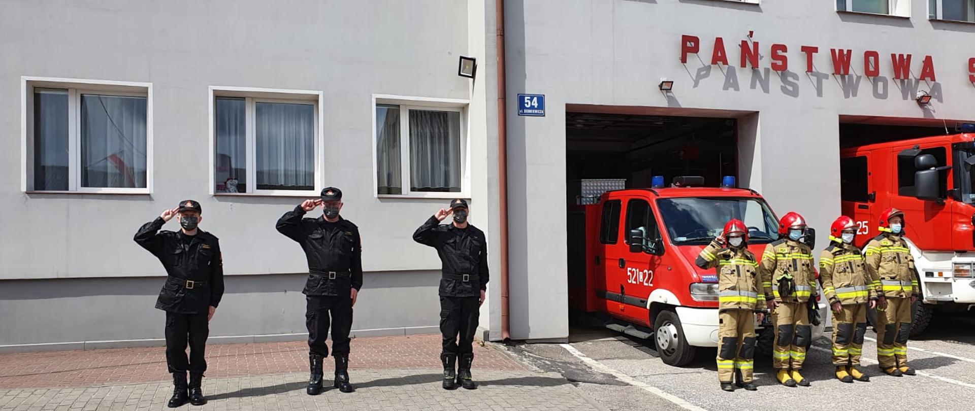 Zdjęcie wykonano przed Komendą Państwowej Straży Pożarnej w Sępólnie Krajeńskim, przedstawia Komendanta Powiatowego Państwowej Straży Pożarnej w Sępólnie Krajeńskim wraz z zastępcą oraz Dowódcą jednostki ubranymi w mundury koszarowe oraz zastęp strażaków w ubraniach specjalnych, stojącymi przed budynkiem. Strażacy stoją na baczność i salutują. 