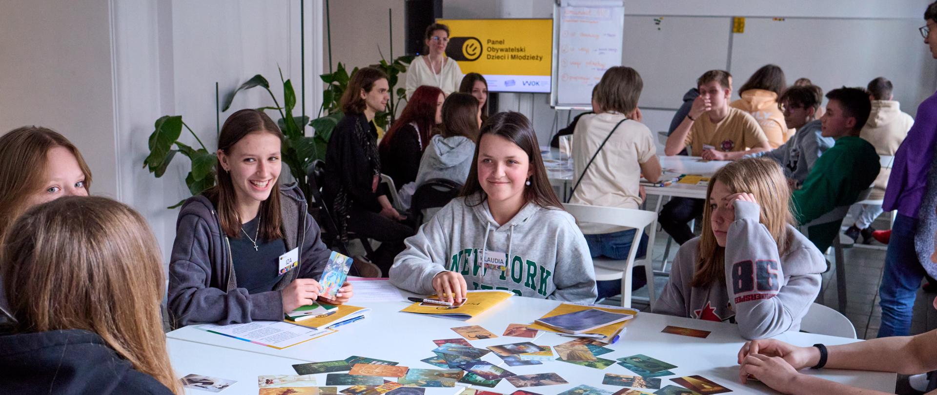 Grupa młodych osób siedzi przy stole z rozłożonymi kartkami edukacyjnymi. W tle inne osoby pracują przy stołach, a na ścianie wisi plakat z napisem „Panel obywatelski dzieci i młodzieży”. Atmosfera jest swobodna i zaangażowana.
