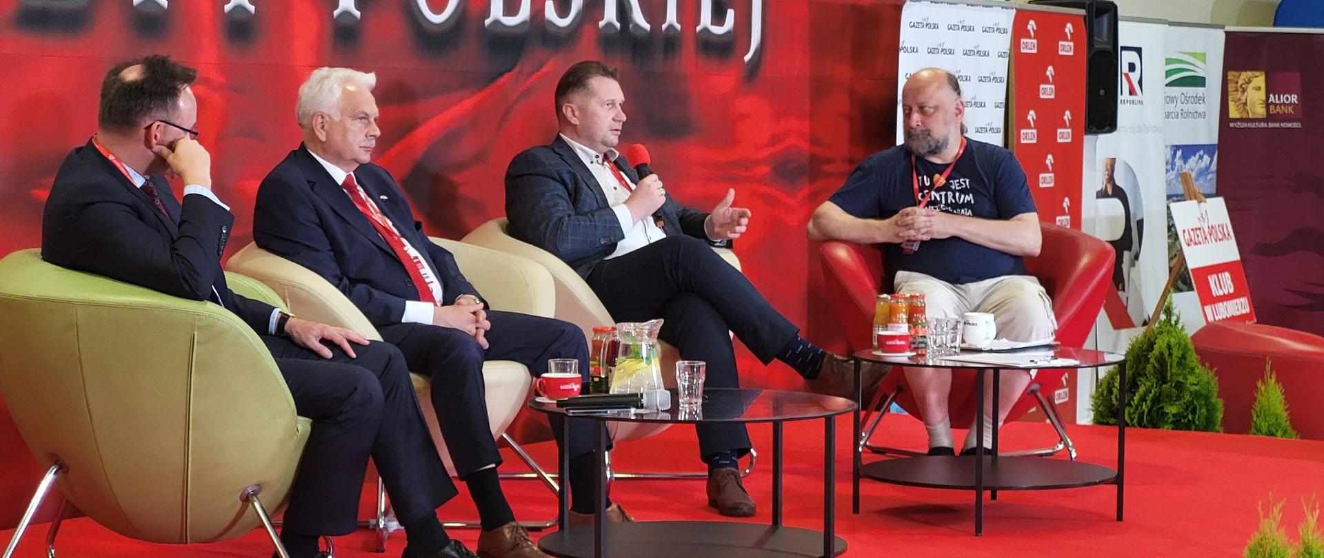 Minister w otoczeniu kilku osób siedzi na fotelu i mówi do mikrofonu, za nimi czerwona ściana.