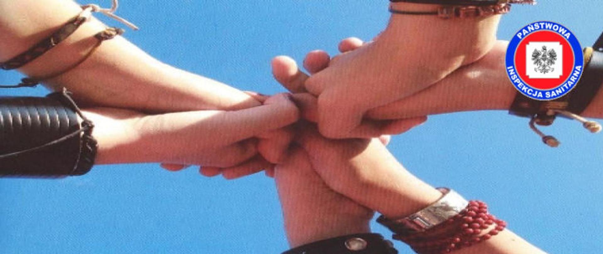 Na tle nieba splecione w uchwycie dłonie sześciu młodych ludzi, z bok ugodło Państwowej Inspekcji Sanitarnej.