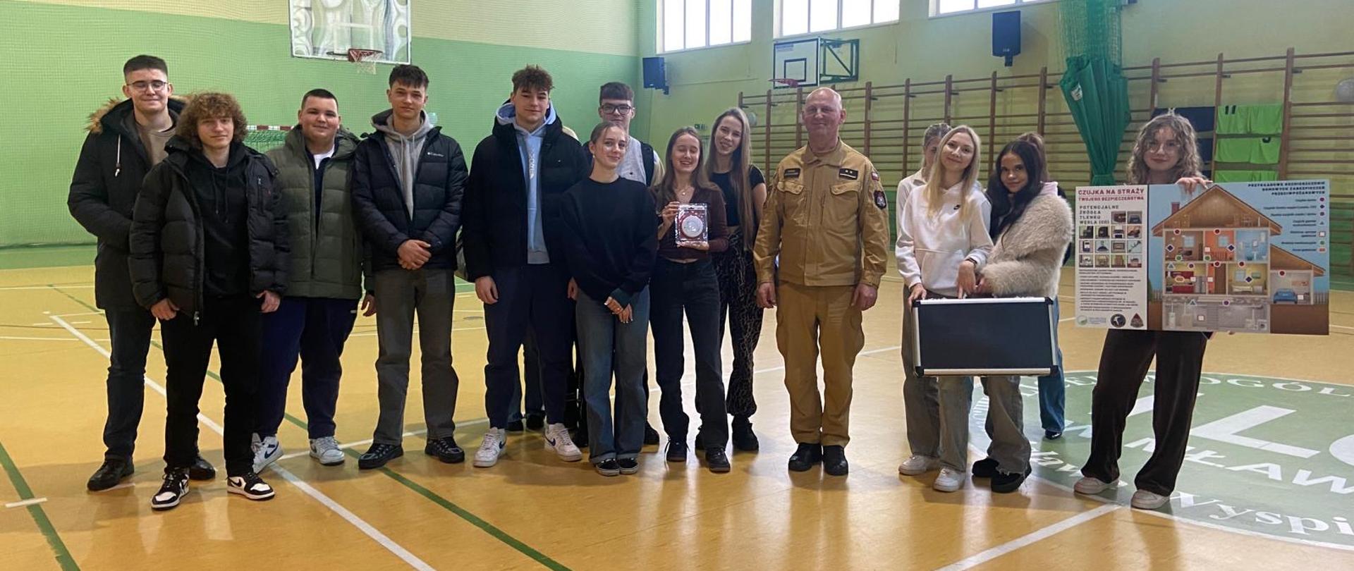 Grupa kilkunastu uczniów i nauczycieli stoi w sali gimnastycznej. W centrum widoczny jest mężczyzna w mundurze strażackim. Część uczniów trzyma plansze edukacyjne i walizkę demonstracyjną. W tle kosze do koszykówki oraz zielone ściany sali.