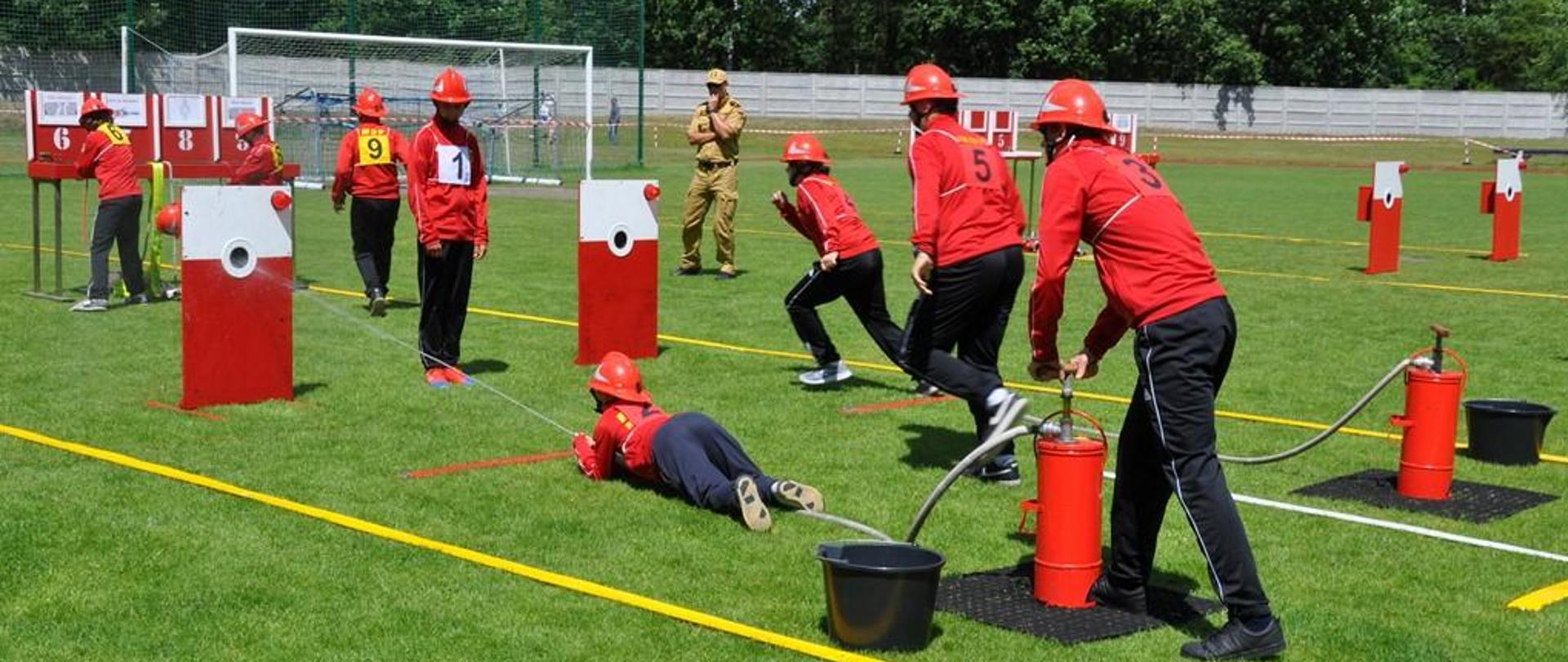 Dzień. Widoka na murawę boiska sportowego, na którym rozstawione są elementy toru do ćwiczenia bojowego. Na torze znajduje się grupka dzieci ubranych w granatowe spodnie dresowe, czerwone bluzy oraz czerwone hełmy. Dzieci wykonują rożne zdania na torze. W tle stoi funkcjonariusz PSP ubrany w mundur służbowy w kolorze piaskowym.