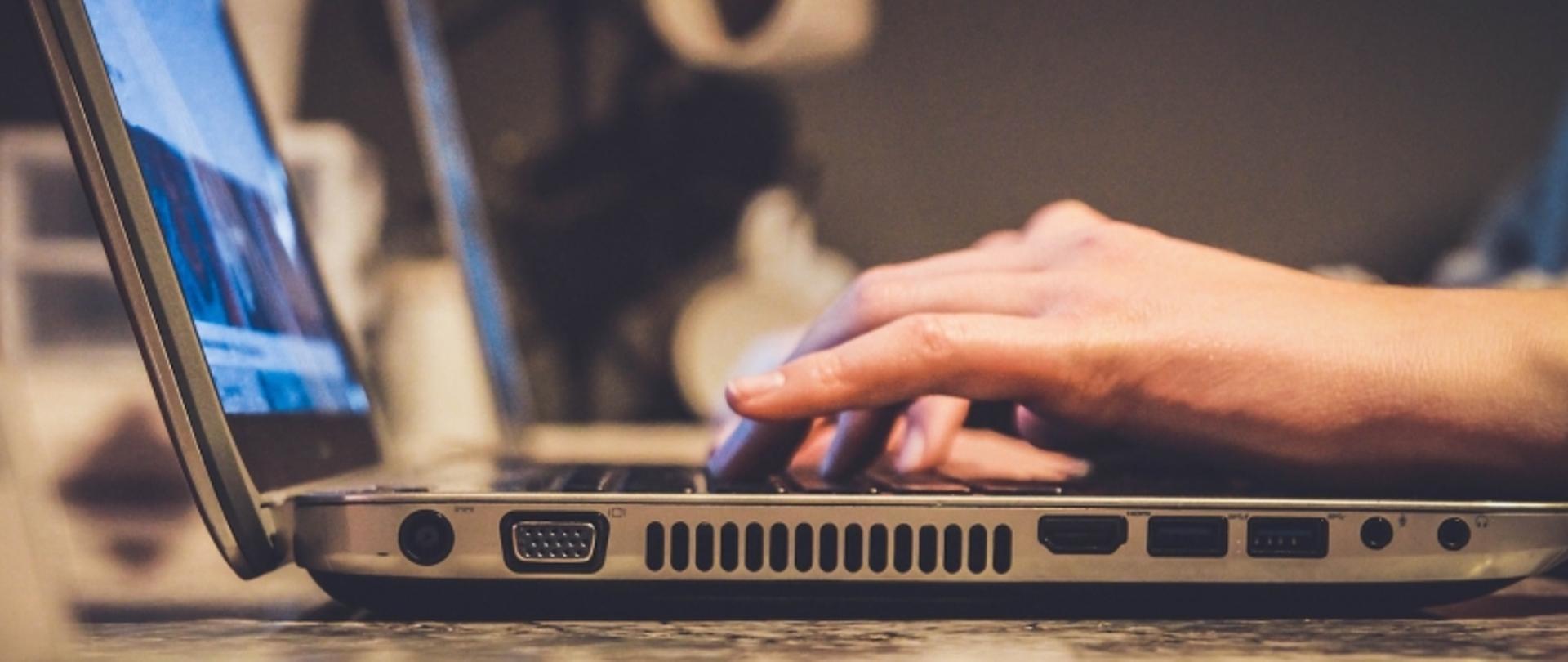 Zbliżenie na dłonie osoby pracującej na laptopie. Widoczne są porty urządzenia, w tym VGA, HDMI, USB i Ethernet, co podkreśla jego funkcjonalność.