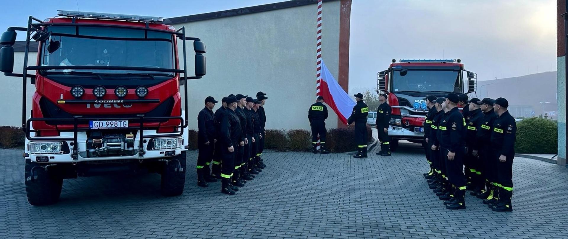 Na zdjęciu widać uroczystą zbiórkę strażaków pod masztem flagowym, poczet flagowy wciąga flagę na maszt, w tle samochody pożarnicze