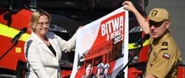 Widok z przodu: Minister klimatu i środowiska Pani Anna Moskwa oraz komendant główny Państwowej Straży Pożarnej gen. brygadier Andrzej Bartkowiak prezentują plakat promujący program "Bitwa o remizy 2023". W tle wozy strażackie. 