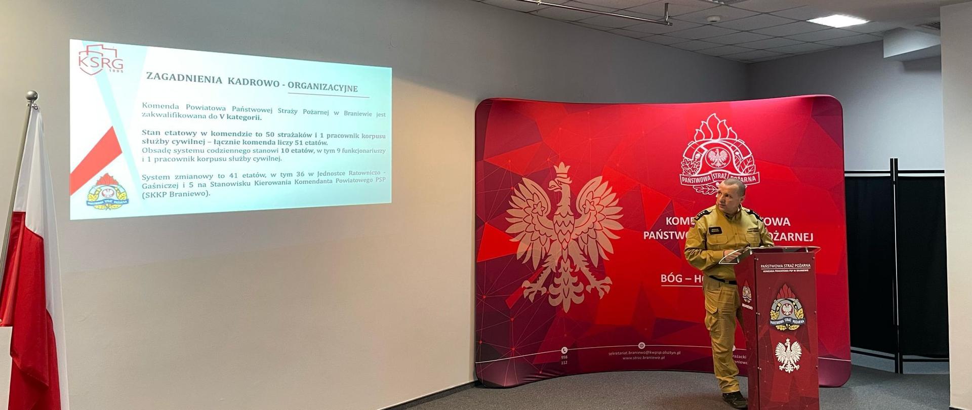 Z prawej strony komendant powiatowy PSP przy mównicy na tle czerwonej ścianki z godłem Polski. Z lewej pokaz slajdów.