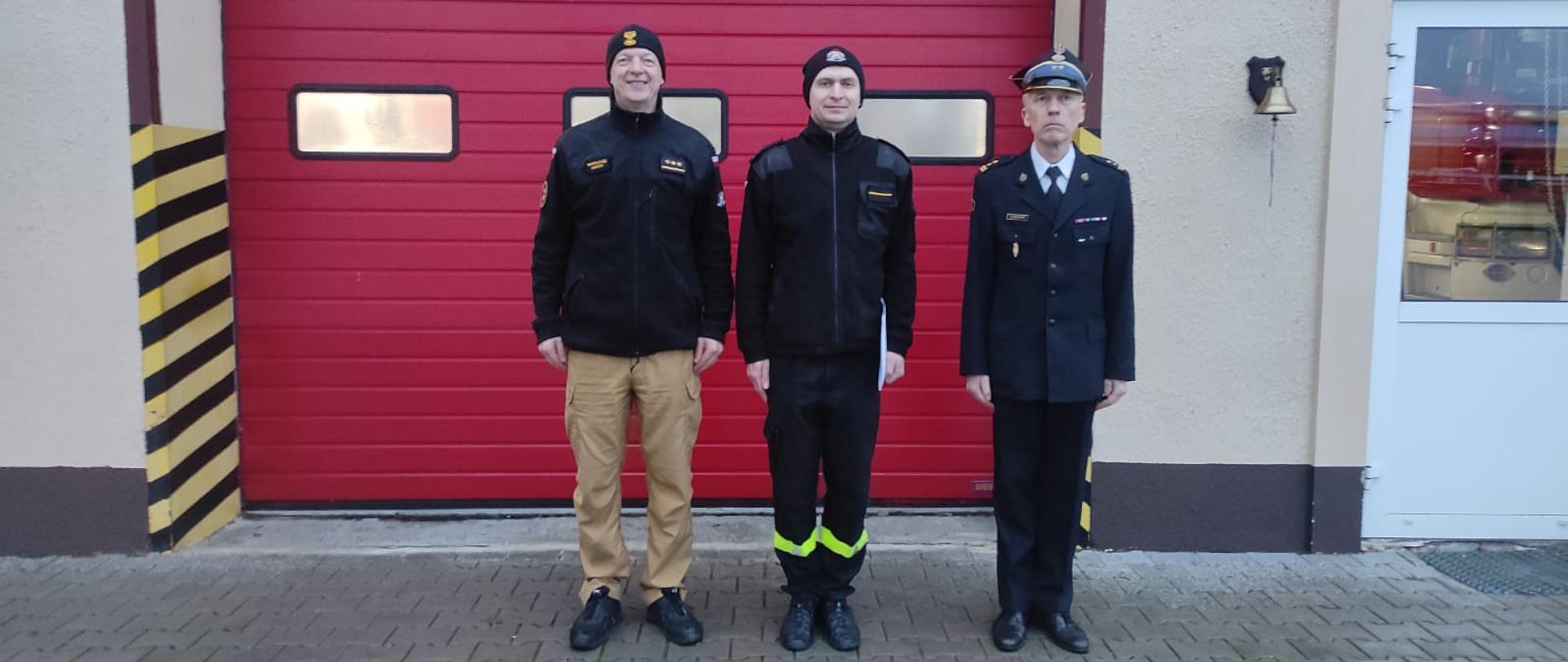 Zdjęcie przedstawia trzech strazaków na tle bramy garazowej po weczeniu awansów.