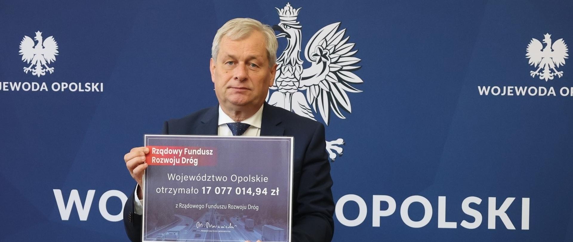 Wojewoda Opolski Sławomir Kłosowski trzyma w rękach symboliczny czek z Rządowego Funduszu Rozwoju Dróg na łączną kwotę ponad 17 mln zł - jest to pula środków przeznaczonych na realizację zadań zwiększających bezpieczeństwo niechronionych uczestników ruchu drogowego (pieszych). W tle niebieska ścianka z godłem państwowym i napisem "Wojewoda Opolski".