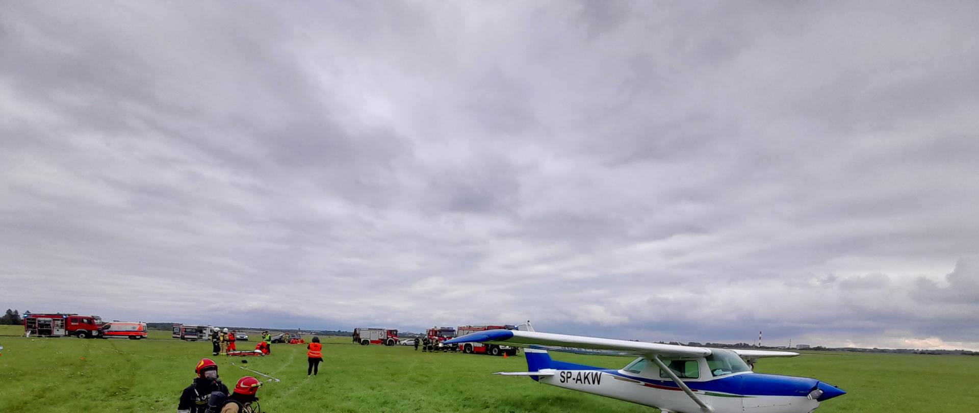 Na zdjęciu widzimy samolot na trawie, strażaków symulujących podawanie piany gaśniczej w tle pojazdy strażackie