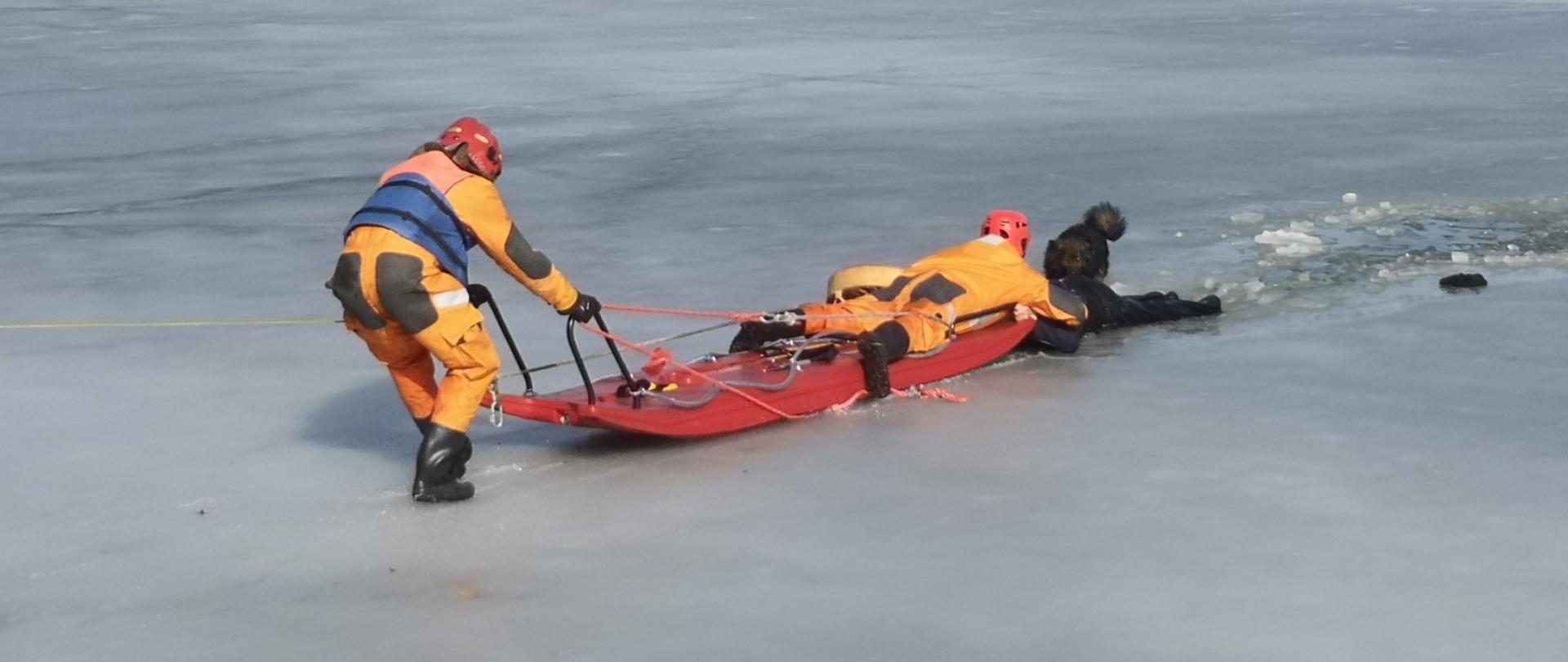 Zdjęcie przedstawia dwóch ratowników PSP z saniami wodno-lodowymi w trakcie ewakuacji mężczyzny z wody.
