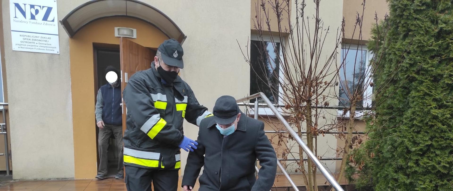 Zdjęcie przedstawia strażaka OSP, który pomaga starszemu mężczyźnie zejść ze schodów. Strażak ubrany jest w ubranie koszarowe i kurtkę ubrania specjalnego. Obaj mężczyźni mają założone maseczki chirurgiczne. Zdjęcie wykonane jest przed budynkiem Niepublicznego Zakładu Opieki Zdrowotnej w Ostrowitem. Z budynku ośrodka zdrowia wychodzi inny mężczyzna. 