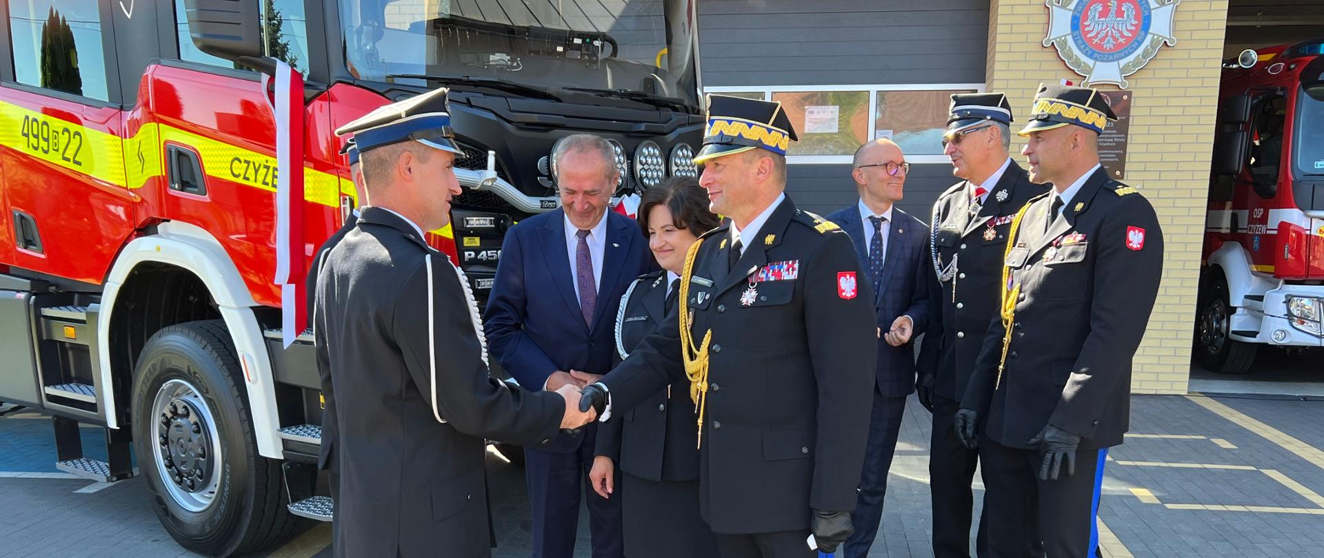 Zastępca komendanta głównego PSP podaje rękę druhowi OSP w geście gratulacji, za nimi grupa strażaków i osoby cywilne oraz nowy samochód rat-gaśn