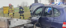 Po lewej stronie widać uszkodzoną przyczepę od samochodu i trzech strażaków, po prawej znajduje się samochód z uszkodzonymi przednimi drzwiami i uniesioną maską, w tle radiowóz Policji