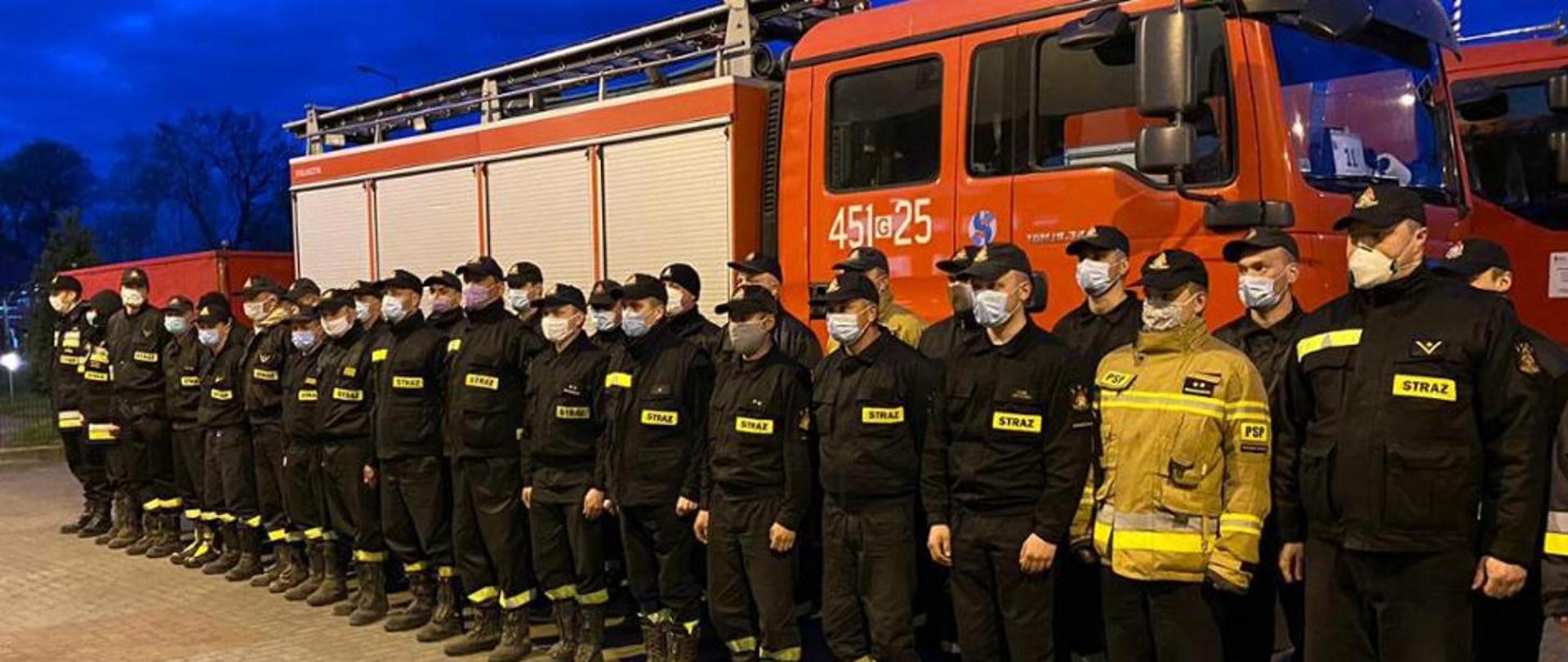 Zdjęcie przedstawia grupę strażaków w mundurach, stojących w dwuszeregu na tle samochodu pożarniczego