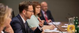 Wiceminister Waldemar Buda oraz trzy inne osoby, siedzi za stołem konferencyjnym i mówi do mikrofonu