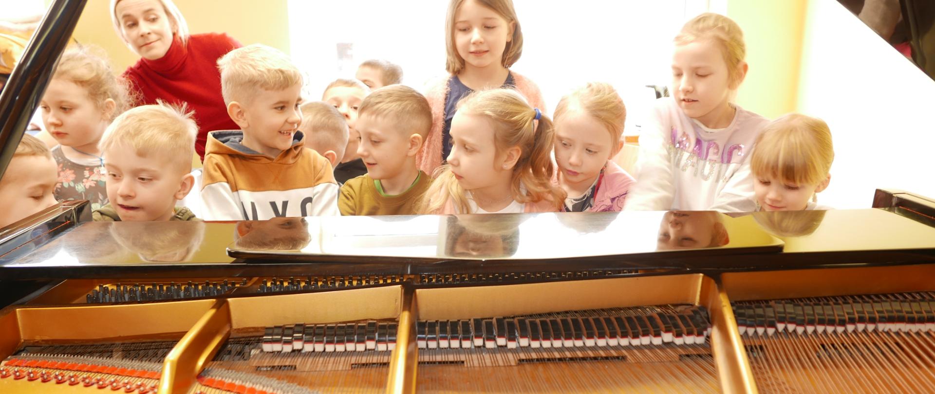 Zdjęcie zrobione z tyłu fortepianu. Klapa jest otwarta i widać struny. Przy klawiaturze siedzi grupa dzieci.