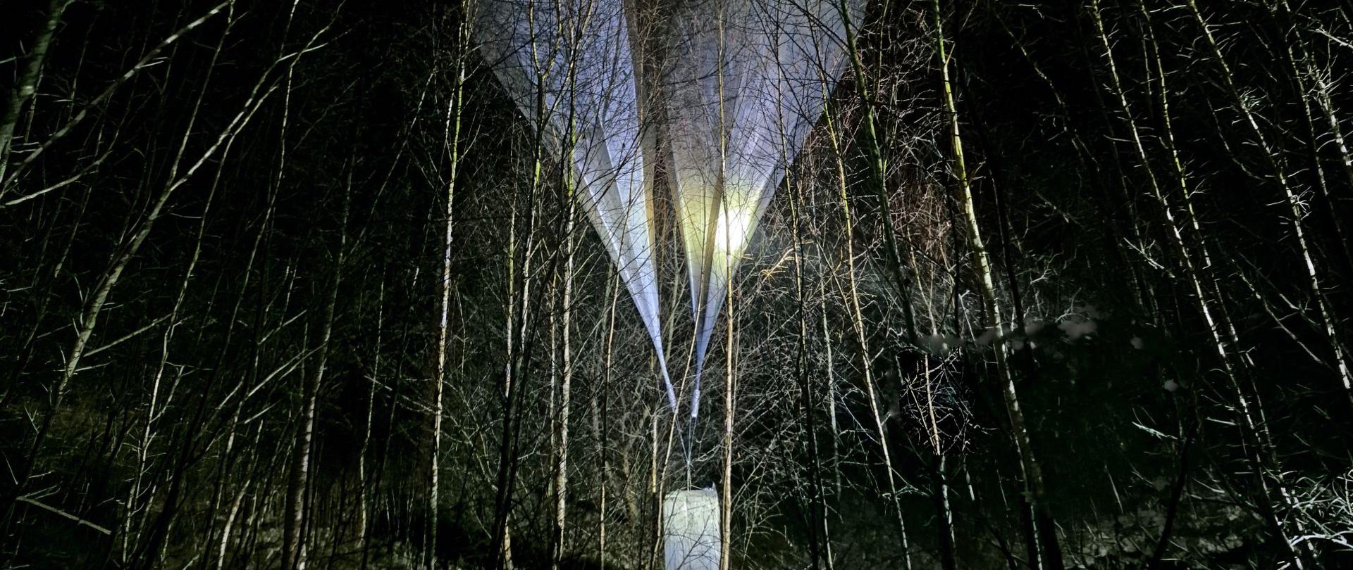 Dwa balony w zalesionym terenie nocą; pod balonami podwieszony pakunek.