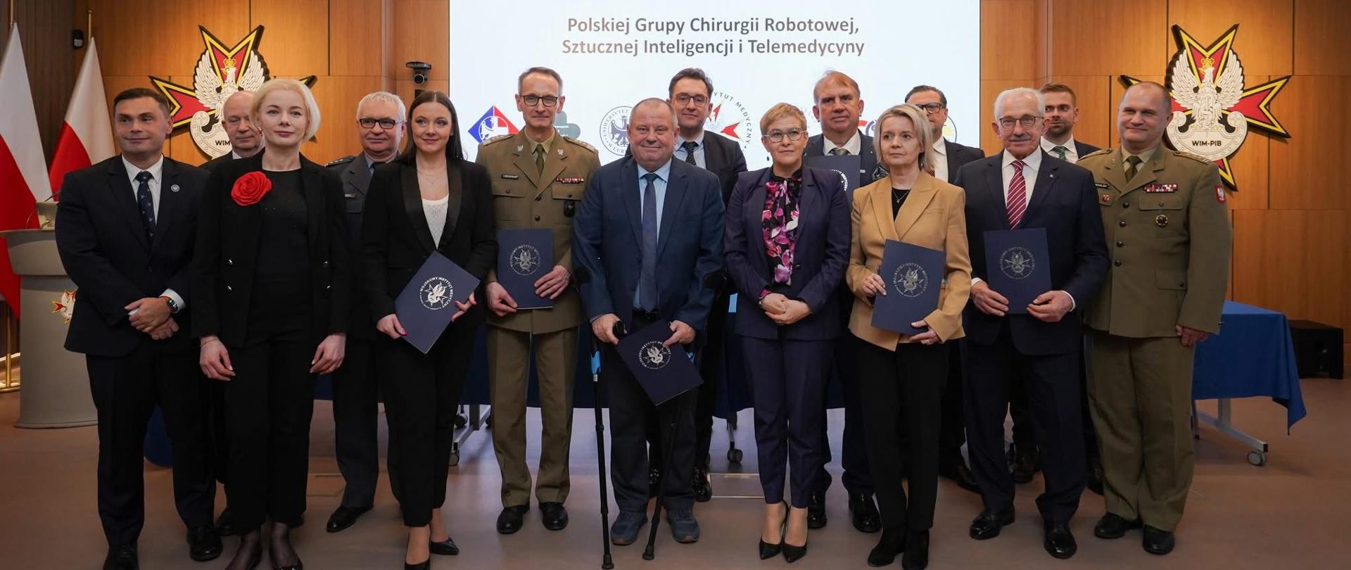 W sali z drewnianymi ścianami stoi grupa kilkunastu osób, pośrodku wiceminister Mrówczyńska, za nimi na ścianie napis Wojskowy Instytut Medyczny.