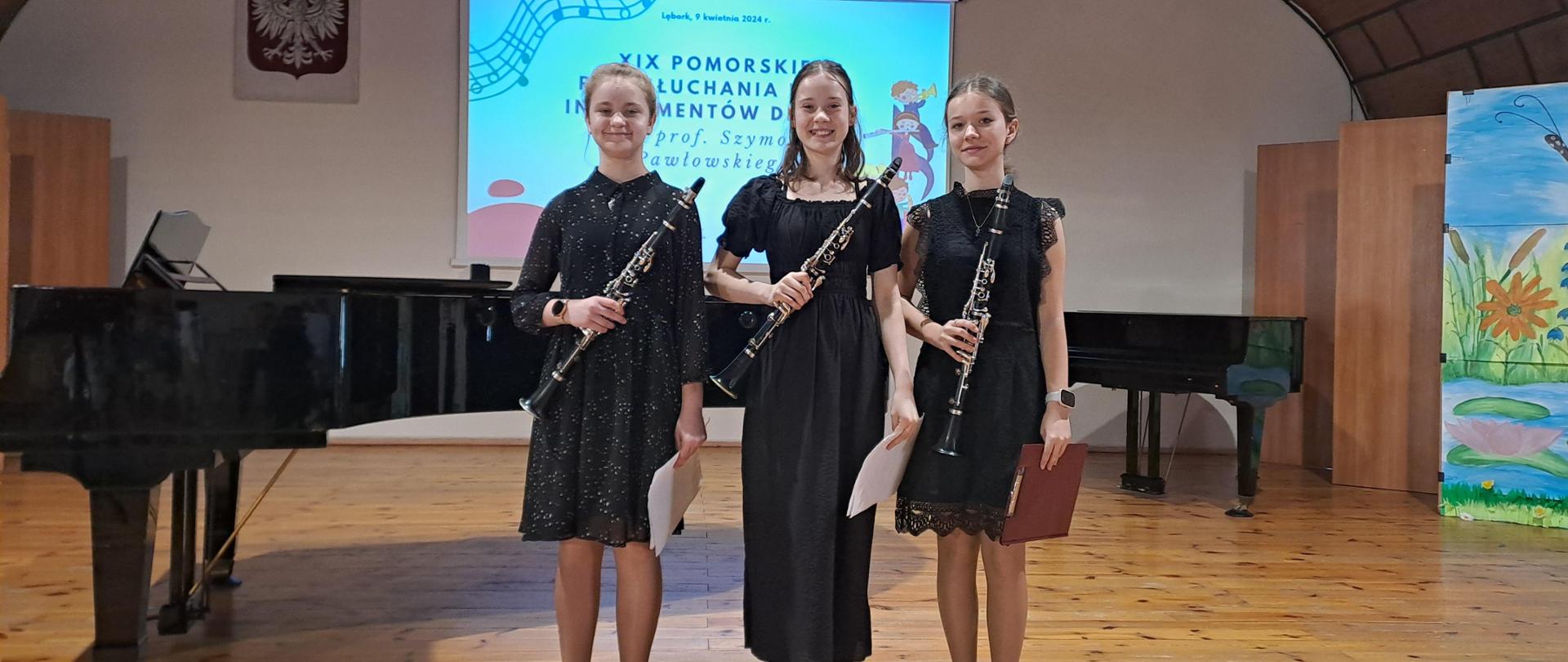 Na sali koncertowej w Lęborku stoją trzy uczennice z klarnetami. W tle fortepian i slajd informujący o lęborskim konkursie.