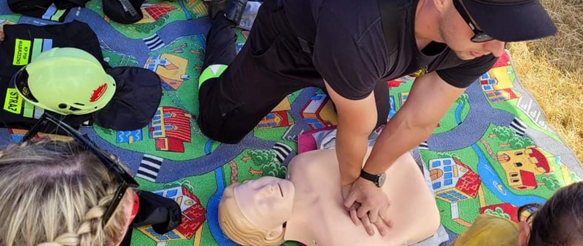 Na zdjęciu strażak udziela instruktażu pierwszej pomocy. W tle plansze edukacyjne. Całość trwa w plenerze pod namiotem. 